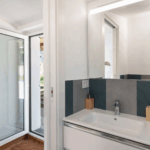 Camera Blu bathroom. Modern white sink, mirror, and door to the outside. Minimalist design.