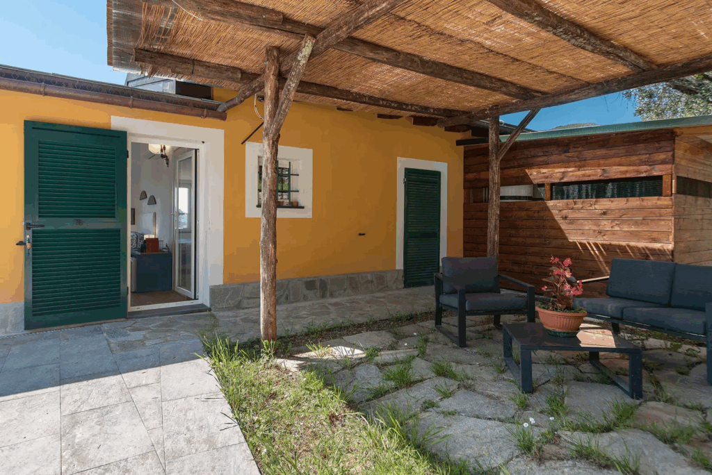 Camera Blu patio. Outdoor seating area with a wooden pergola and stone flooring. Yellow building with green shutters.
