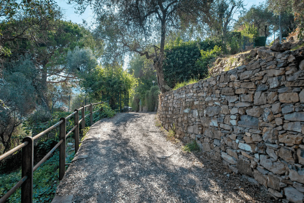 Casa di Pietra pathway. Stone wall lines a sun-dappled path through lush greenery. Railing on the left.