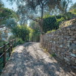 Casa di Pietra pathway. Stone wall lines a sun-dappled path through lush greenery. Railing on the left.