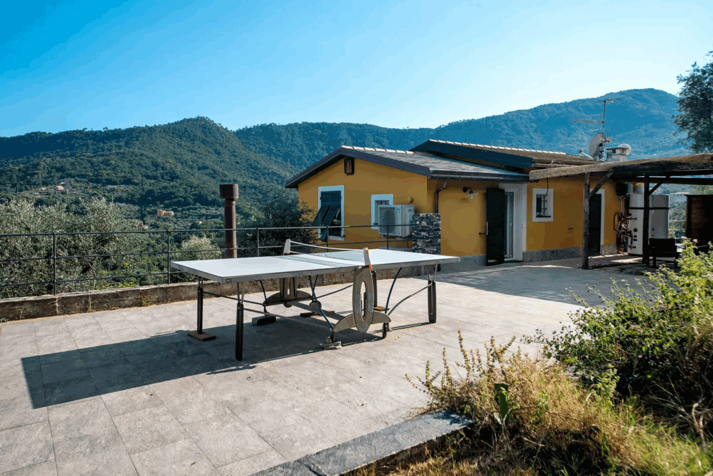 Casa di Pietra terrace view. Ping pong table on patio with mountain backdrop. Yellow building with blue shutters.